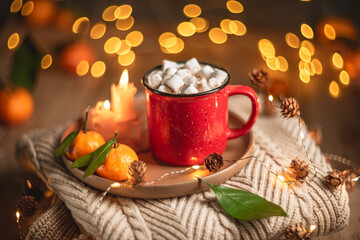 Red mug filled with hot chocolate and marshmallows resting on a tray with tangerines, surrounded by warm knit fabric and glowing christmas lights, creating a cozy winter scene