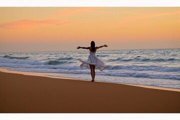 Fototapeta premium Young Woman in a Flowing White Dress Embracing the Sunset on a Serene Beach