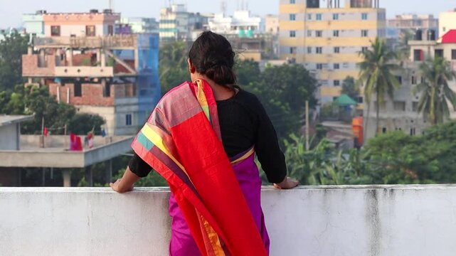 A Bengali woman in a saree stands alone on a rooftop, gazing at the beautiful outdoor scenery. Her saree veil sways gently in the breeze. Back view.
