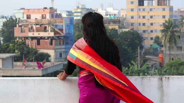 A Bengali woman wearing saree standing alone on the rooftop, gazing out at the cityscape. Back view.