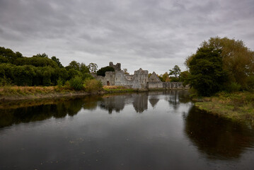 Fototapeta premium Desmond Castle in Adare, Ireland, reflected in a tranquil river surrounded by lush greenery and a cloudy sky