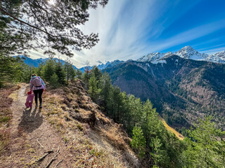 Fototapeta premium Mother walking with toddler on scenic hiking trail looking at snow capped mountain peaks of Julian Alps seen from top of Monte Nebria Cima Est, Friuli Venezia Giulia, Italy. Wanderlust in early spring