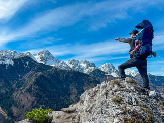 Father with baby carrier looking at snow capped mountain peaks of Julian Alps seen from top of Monte Nebria, Friuli Venezia Giulia, Italy. Wanderlust in alpine wilderness in spring. Positive emotions