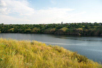 Fototapeta premium Scenic view of a tranquil river with lush green hills and a small village in the background under a clear blue sky. Perfect for nature, travel, and landscape themes