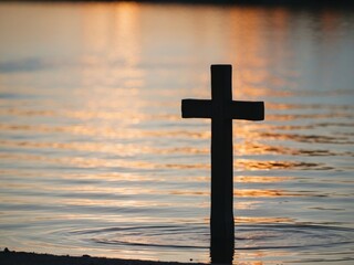 Silhouette of wooden Christian cross on reflective water lake river during sunset.