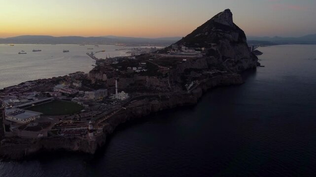 Aerial view of Gibraltar at sunset. Street lights on. Europa Sports Complex Stadium. Famous travel destination. Bay of Gibraltar. City of Gibraltar and La Linea, in background. Drone going backwards.