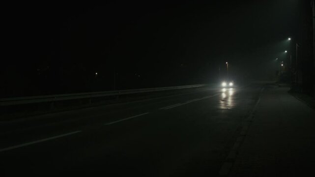Dark street at night with single car passing by on the road, headlights and streetlights glowing in the dark, illuminating the night. Lonely car. Melancholy. High quality 4k footage