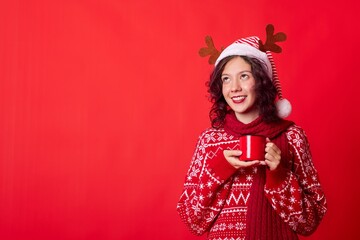 Smiling young woman in a red sweater and Santa hat holding a red mug of hot cocoa on red background
