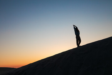 Silhouette of a woman walking on a sand dune at sunset, under a tranquil and colorful evening sky....
