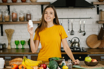 Young attractive happy young woman standing at the kitchen table and drinking dietary supplements,...