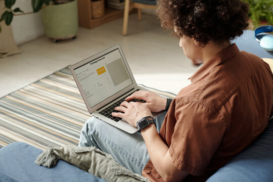 Young modern man in casualwear sitting on couch in home environment and using AI image generator on laptop