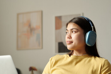 Young serene woman in headphones looking aside while sitting in front of camera in domestic room and listening to music