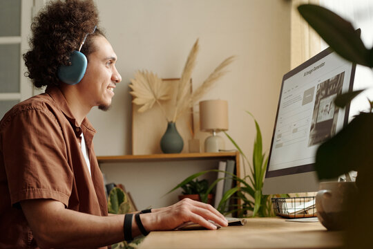 Smiling young man listening to music in headphones and looking at screen of desktop computer during work