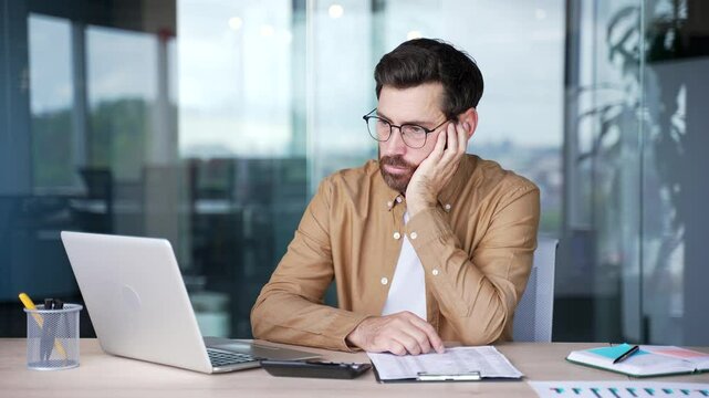 Upset bearded businessman is bored sitting at workplace at desk in business office. Exhausted man is tired from working with laptop and doing paper work. A lazy worker procrastinates, gets distracted