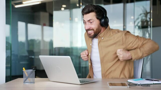 Excited businessman in wireless headphones watching sports match or competition using laptop while sitting in office. Happy man cheering for bids at auction, celebrating success showing achievement