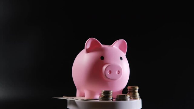 Hand of male inserts a banknote into a pink piggy bank that sits on a rotating pedestal, surrounded by scattered coins and additional notes, set against a dark background