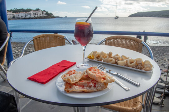 un verre de sangria avec des tapas, du pain grill&eacute; &agrave; la tomate et des calamars frits, sur une petite table en ext&eacute;rieur face &agrave; la mer