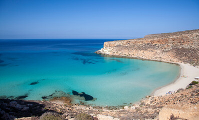 A breathtaking view of Spiaggia dei Conigli in Lampedusa, featuring crystal-clear turquoise waters, white sand, and rugged cliffs under a bright blue sky.