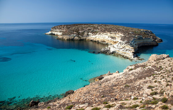 A breathtaking view of Spiaggia dei Conigli in Lampedusa, featuring crystal-clear turquoise waters, white sand, and rugged cliffs under a bright blue sky.