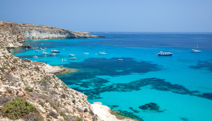 A stunning view of Lampedusa's southern coast, showcasing turquoise waters, anchored boats, dramatic cliffs, and a serene Mediterranean ambiance under a clear blue sky.