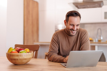 Portrait of handsome freelancer smiling and working online over laptop while sitting at dining table