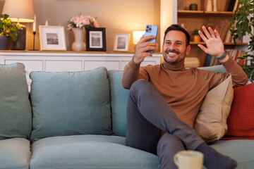 Portrait of cheerful businessman waving hand and greeting while talking over video call on sofa at...
