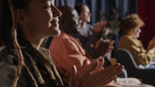 Side tilt closeup of Caucasian female spectator in foreground watching stand-up comedy performance and clapping happily along with other blurred silhouettes of spectators in background