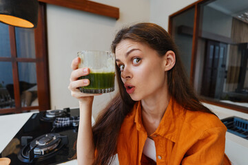 Young woman holding a green smoothie in a modern kitchen, expressing excitement about healthy...