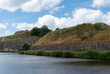 Obraz premium A tranquil river flows beside ancient fortifications covered in grass and shrubs. A house rests atop the hill, framed by bright clouds and blue sky, creating a picturesque setting