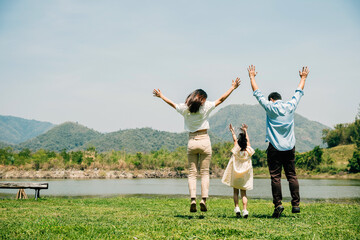 Asian family father mother and daughter jumping front lake with raised hands sunny day in park,...