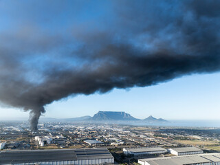 Fototapeta premium Factory fire spilling huge smoke cloud into Cape town sky aerial view with Table Mountain in background