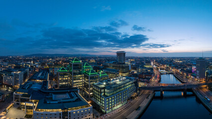 A dusk view of Dublin's modern skyline with the iconic green-lit Bank of Ireland building, a river reflecting the city lights, and a bridge crossing over it.