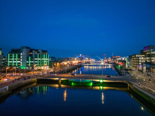A dusk view of Dublin's modern skyline with the iconic green-lit Bank of Ireland building, a river reflecting the city lights, and a bridge crossing over it.