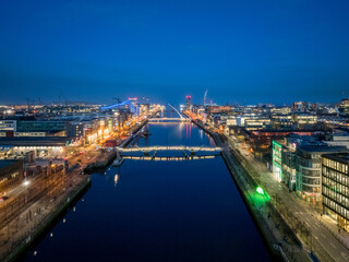 A dusk view of Dublin's modern skyline with the iconic green-lit Bank of Ireland building, a river reflecting the city lights, and a bridge crossing over it.