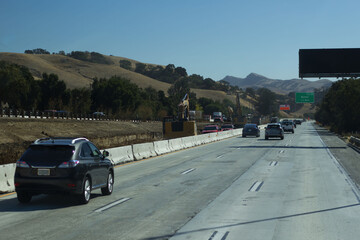 Traffic flows past construction on a highway surrounded by hills on a clear day