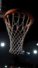 A basketball net is lit up in the dark. The net is white and orange. The lights are on in the background