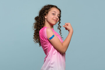 Girl flexing arm with a blue bandage after vaccination, wearing a pink shirt. Healthcare safety
