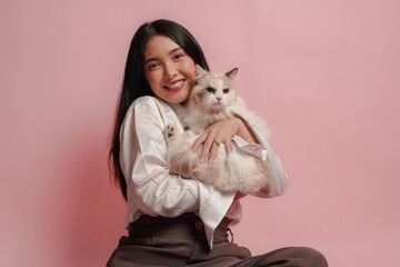 Pretty young Asian woman in white shirt is holding a white ragdoll cat and smiling to the camera.
