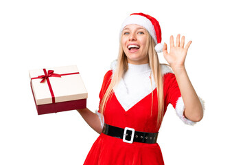 Young blonde woman with christmas hat and holding a gift over isolated chroma key background saluting with hand with happy expression