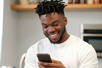 Young man with stylish hairstyle smiles while using smartphone in a bright kitchen, enjoying technology, relaxation, and modern lifestyle in casual attire