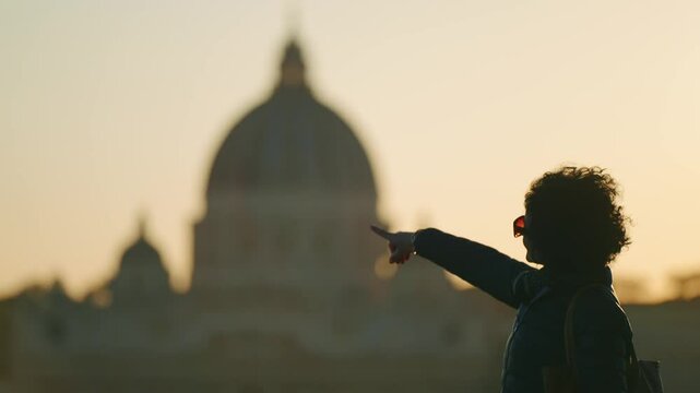 A woman tourist with curly hair points towards the iconic dome of St. Peter's Basilica in Rome during a beautiful sunset.