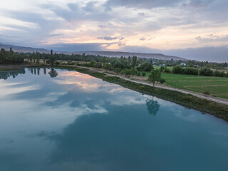 Lake at sunset, trees and grass on the shore, clouds in the sky. Drone view of the summer landscape.