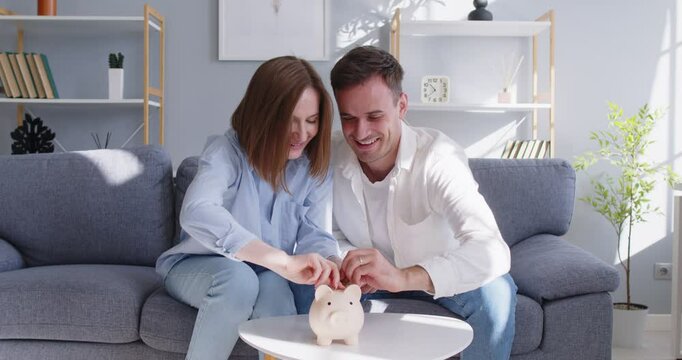 Happy young married couple at home putting coins in piggy bank, smiling spouses saving money, planning family budget, making investment in future, thinking of payment, personal rational economy