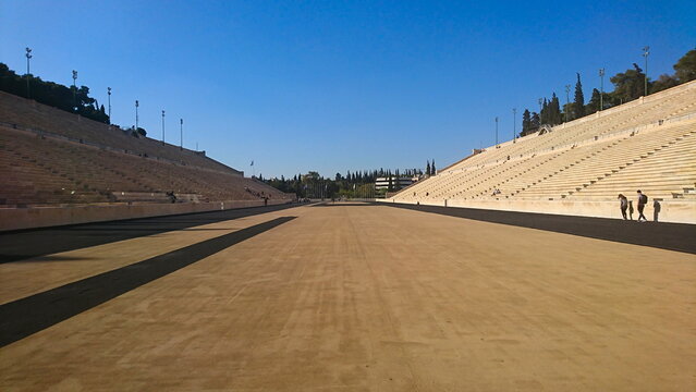 Athens, Greece - 29.3.2018: Tourists walking along the track on the field of Panathenaic Stadium with marble steps on both sides and flags at the end under a clear blue sky in the summer before Covid