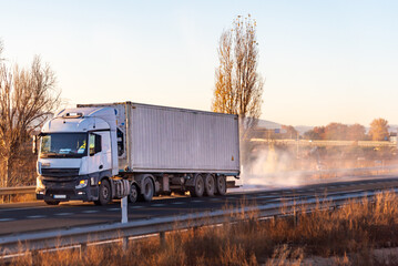 Truck with a shipping container driving on a highway, with the semi-trailer emitting a cloud of smoke after a tire on the rear axle burst.