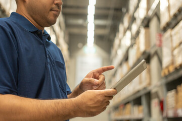 Warehouse worker wearing a hat and blue shirt hands holding tablet check stock on tall shelves in...