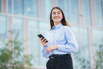 Fototapeta premium Young woman smiles while holding a smartphone in an urban setting with modern architecture in the background