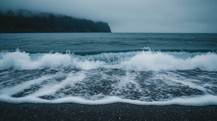 Obraz premium Foamy waves crashing on a dark beach, with a misty mountain range in the distance.