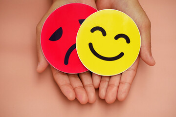 Hand holding round emoji card, smiling face and sad face behind happy face. Concept of split...