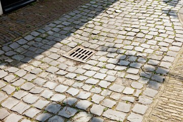 Fototapeta premium Cobblestone pavement with a metal drain cover. Small plants and debris are scattered. Sunlight casts shadows.